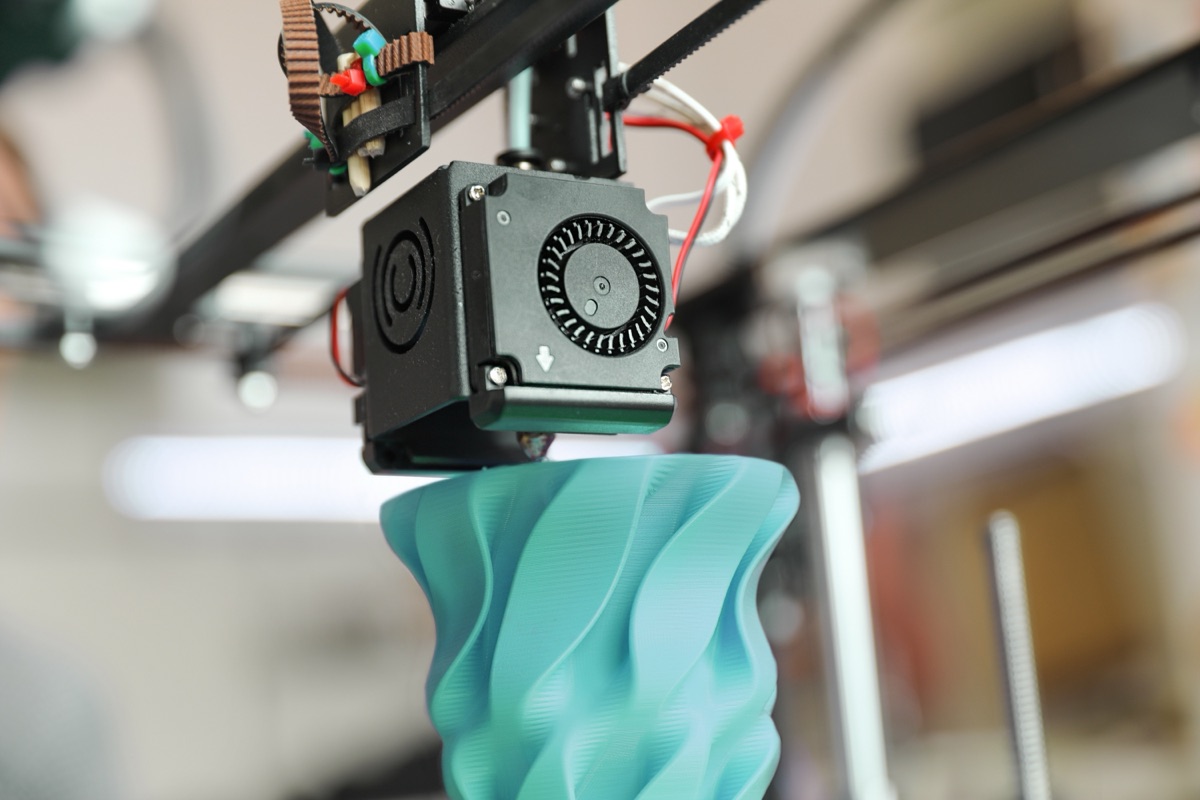 Close-up of a print head extruding a teal sculptural form with a twisted wave profile