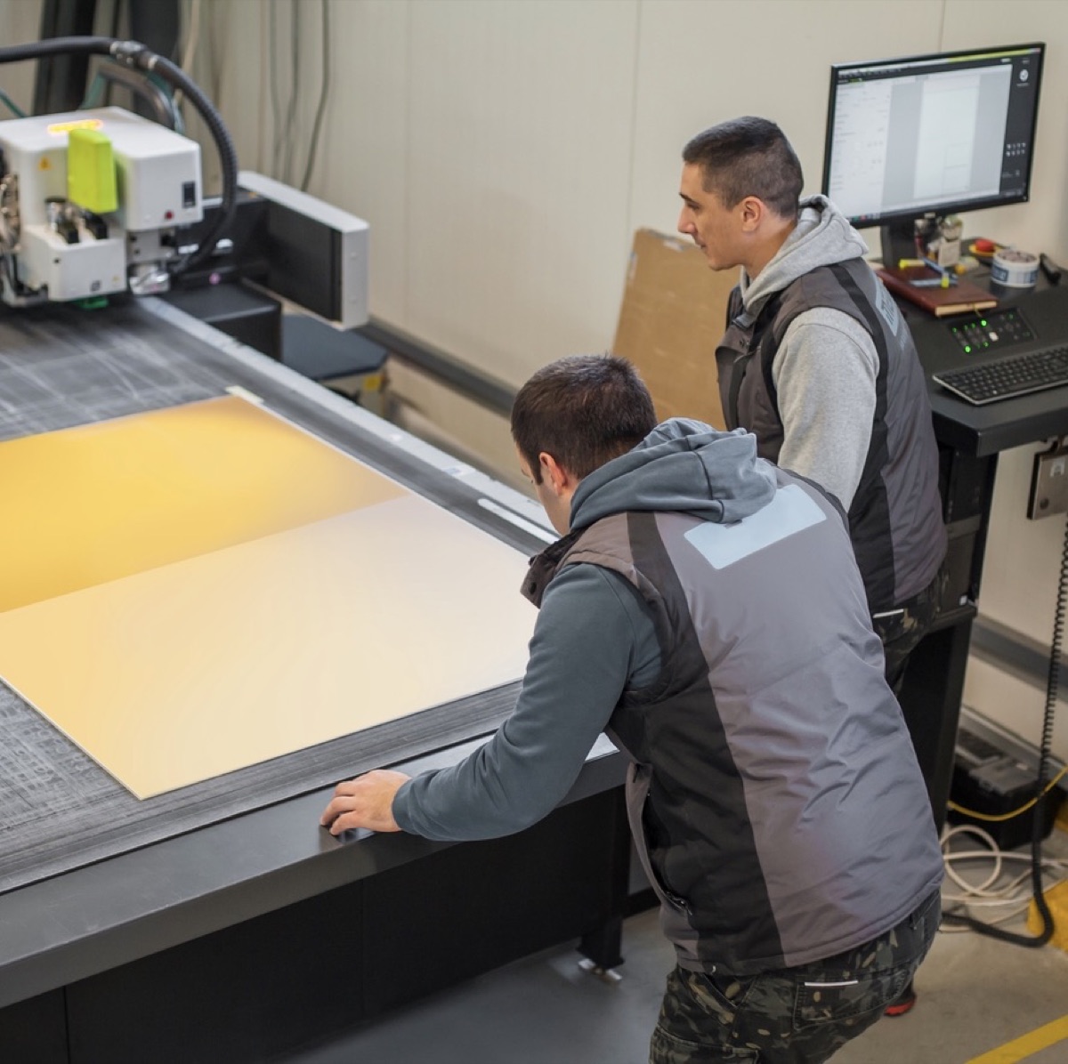 Two operators checking a sheet on the flatbed cutting table against the cut program on screen