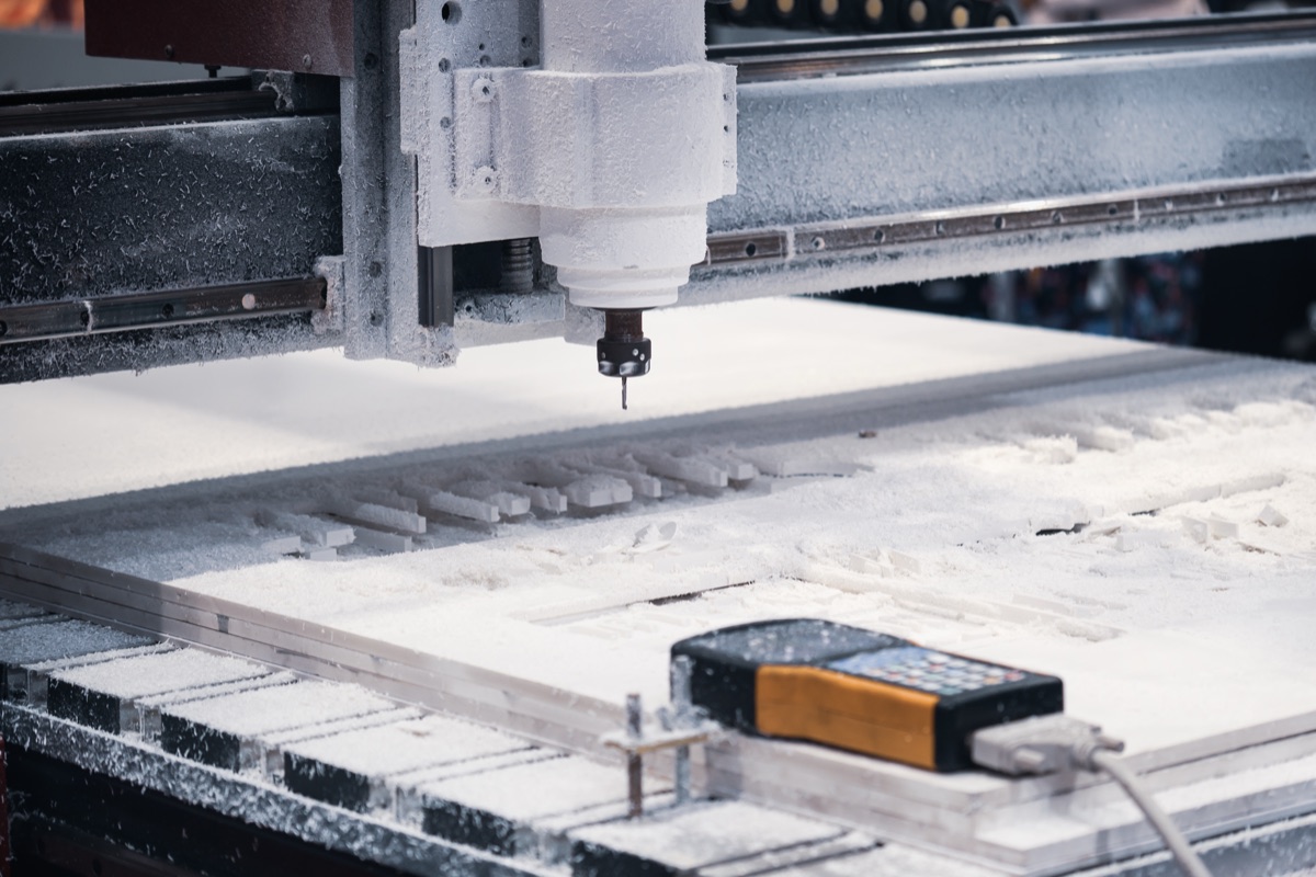 Router gantry over a white sheet of machined components, pendant controller resting on the bed