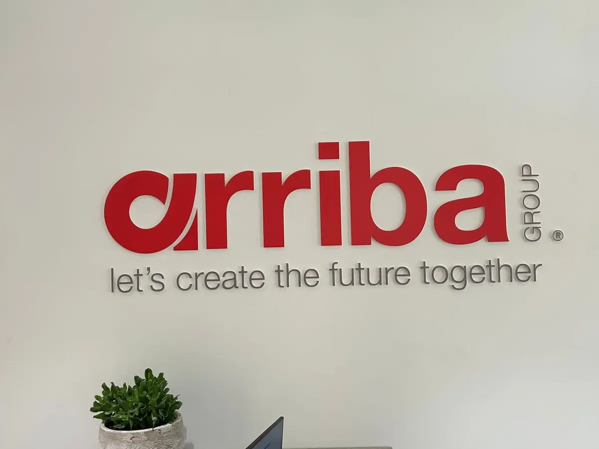 Dimensional acrylic Arriba Group logo in red with a grey tagline installed on a white reception wall above a potted plant