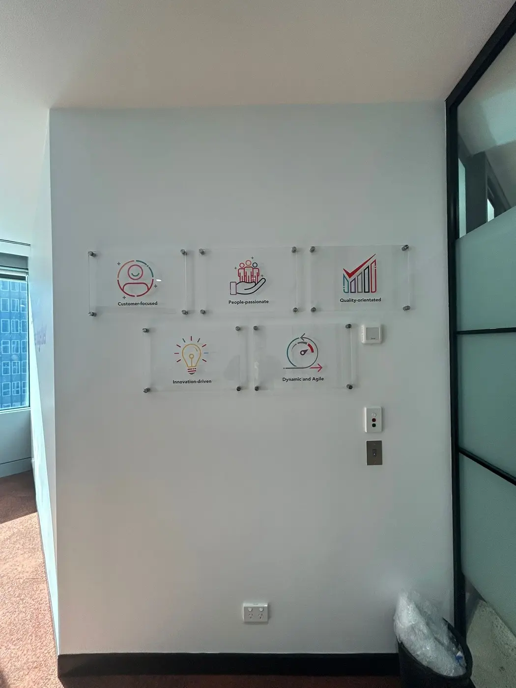 Five clear acrylic panels carrying the Arriba Group values as brand-coloured icons, mounted on silver barrel standoffs across a white office wall