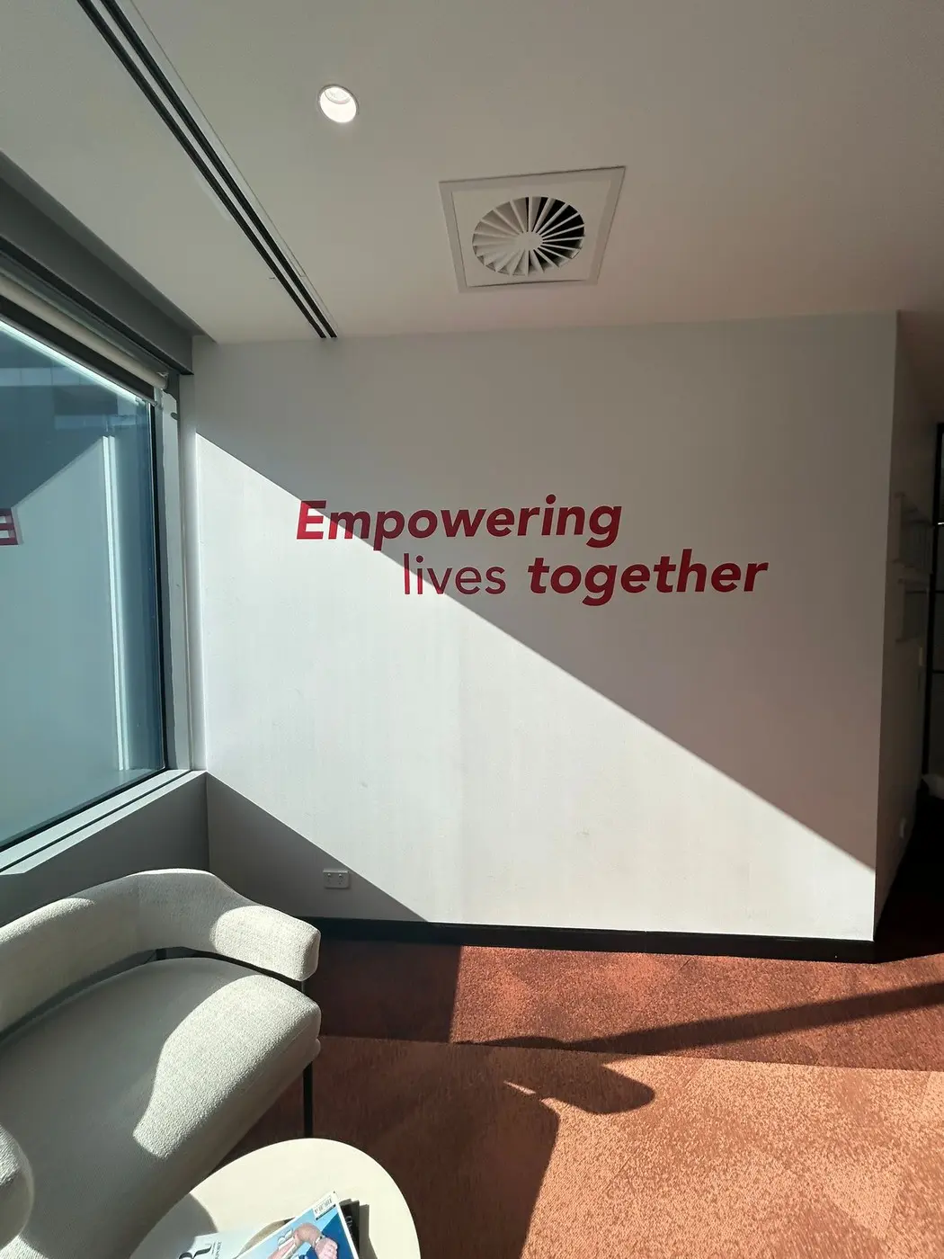 Dark red cut vinyl wall lettering reading Empowering lives together on a white wall beside a window in the Arriba Group office lounge
