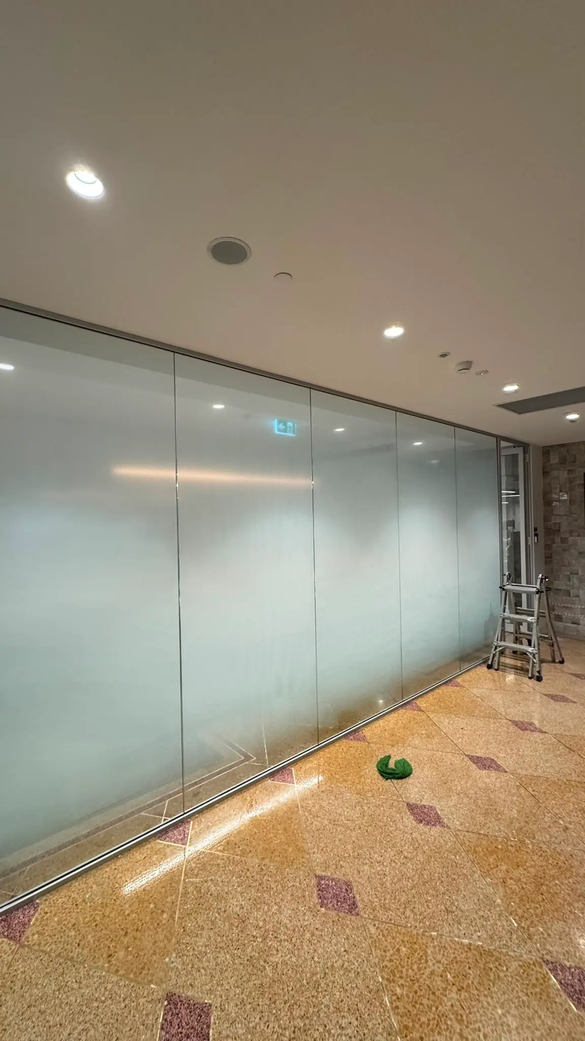 Full height glazed partition beside a terrazzo floored corridor with privacy film applied across every pane and a step ladder standing at the far end