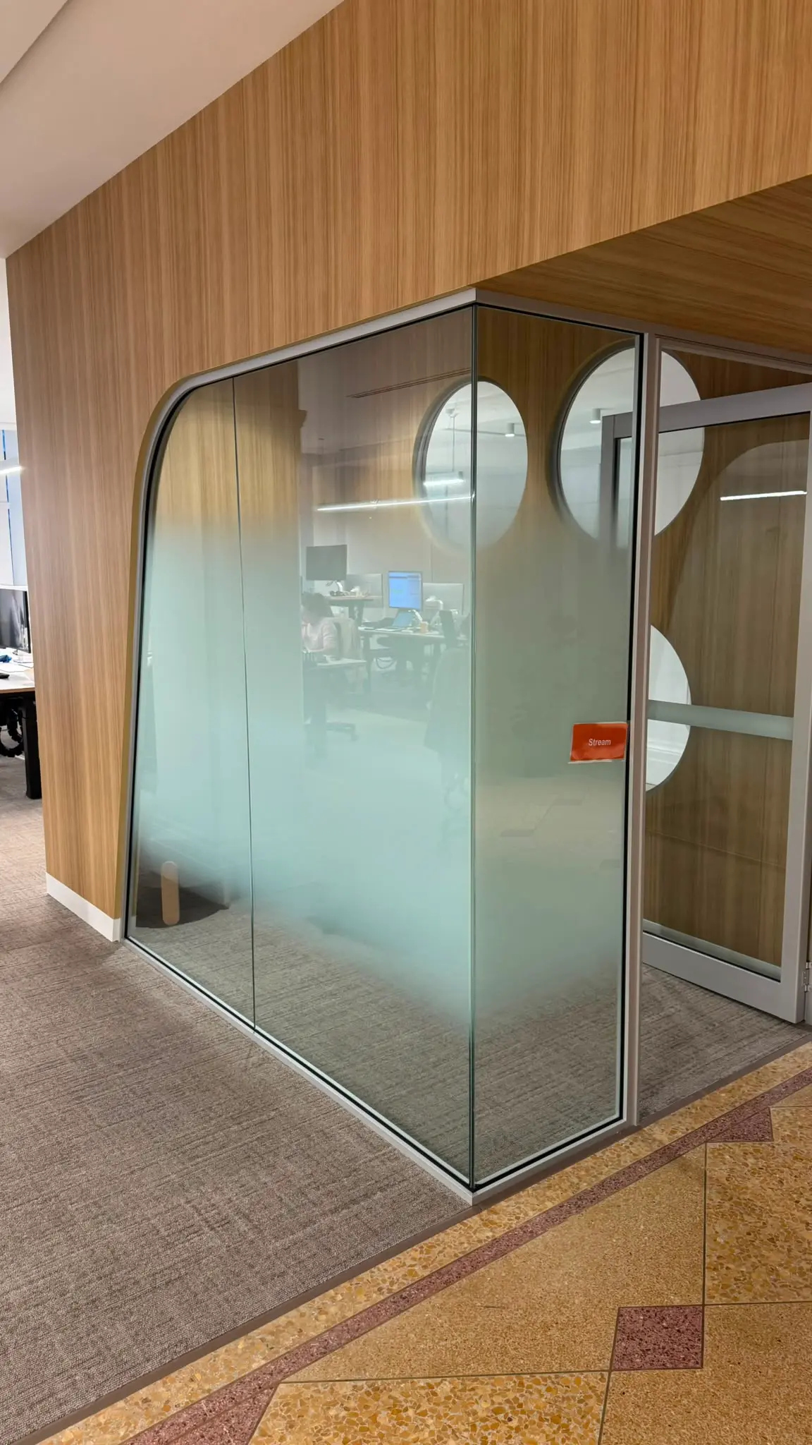 Curved corner glass meeting pod with the printed gradient across its lower glass and large pale circles on the clear glazing above, beside an orange room name label reading Stream