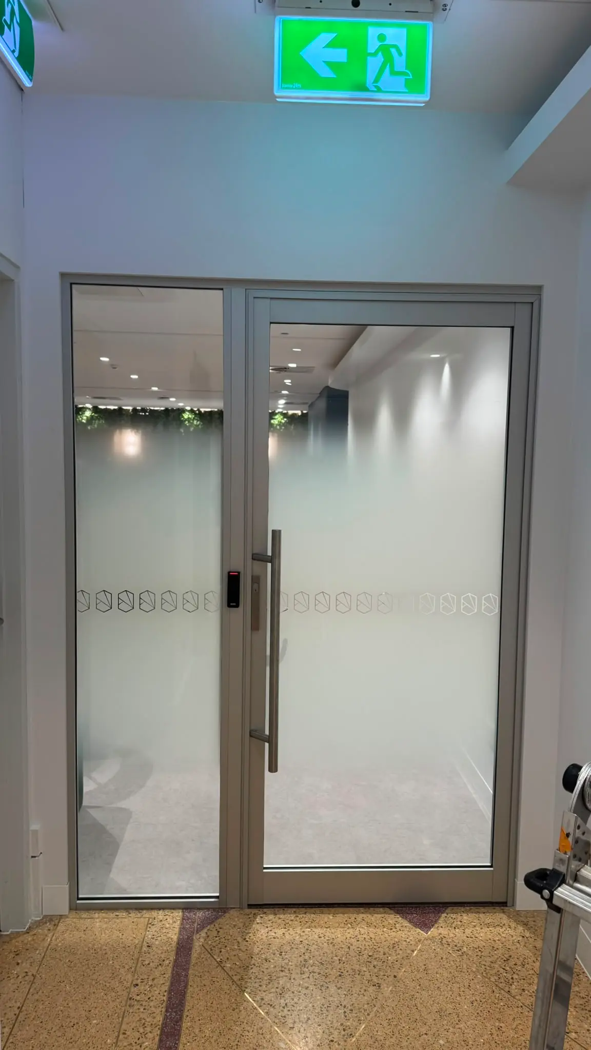 Second glazed entrance door seen from the terrazzo lift lobby with gradient film, an exit sign above and planting visible inside