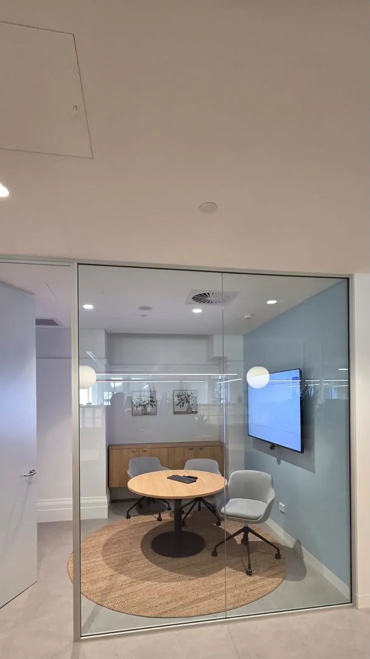 Internal glazed meeting room with completely clear glass showing a round table, three grey chairs, a timber credenza and a wall mounted screen