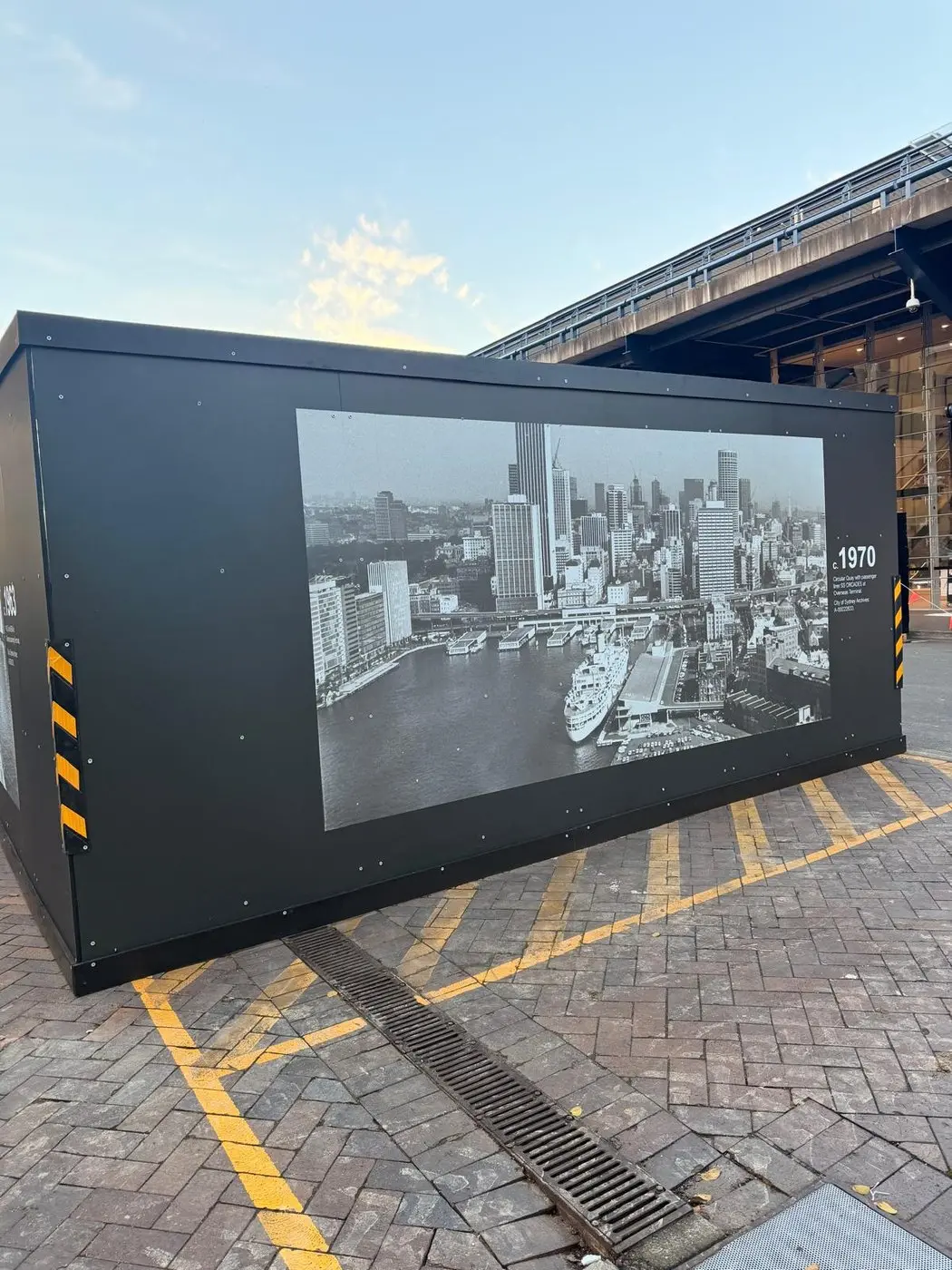 End wall of the site compound hoarding printed with a 1970 aerial photograph of Circular Quay, chevron markings on both corners