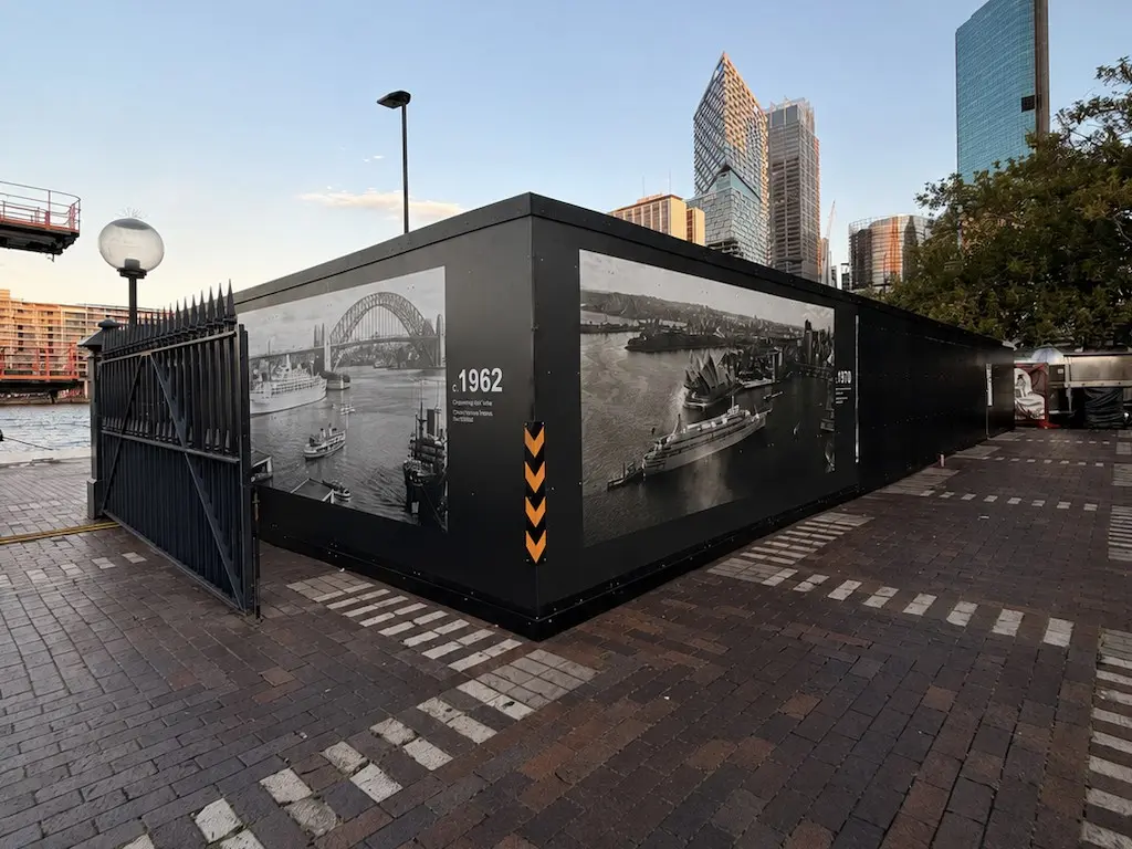 Site compound hoarding on the Circular Quay forecourt wrapped around its corner with c.1962 and 1970 archival harbour photographs, heritage gates alongside and the city skyline behind