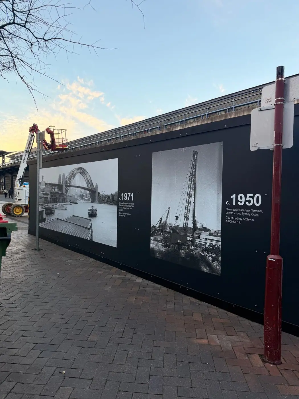 Compound hoarding on the terminal forecourt carrying 1971 and 1950 archival panels, with an elevated work platform alongside during install