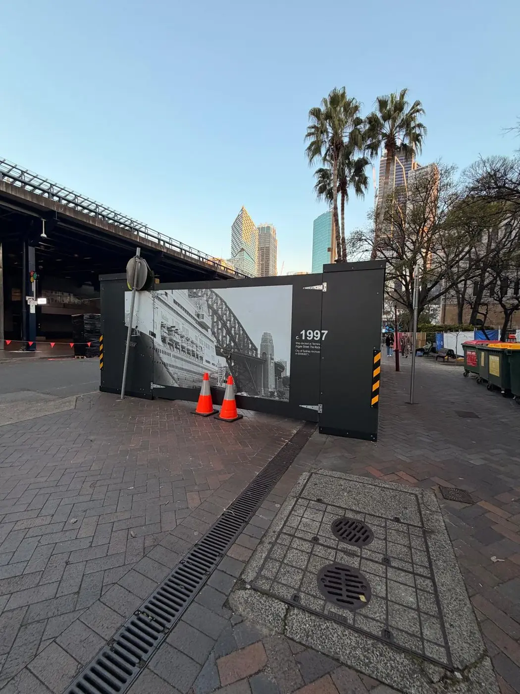 Hoarding gate leaves at the terminal entry wrapped in a 1997 archival liner photograph, traffic cones set out in front
