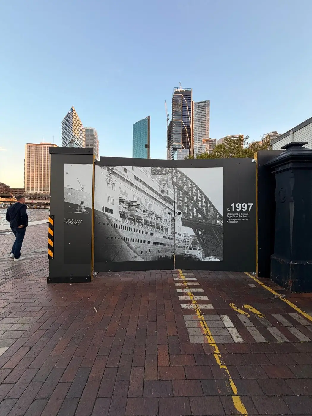 Pedestrian walking past a hoarding gate panel printed with a 1997 photograph of a liner berthed beneath the Harbour Bridge