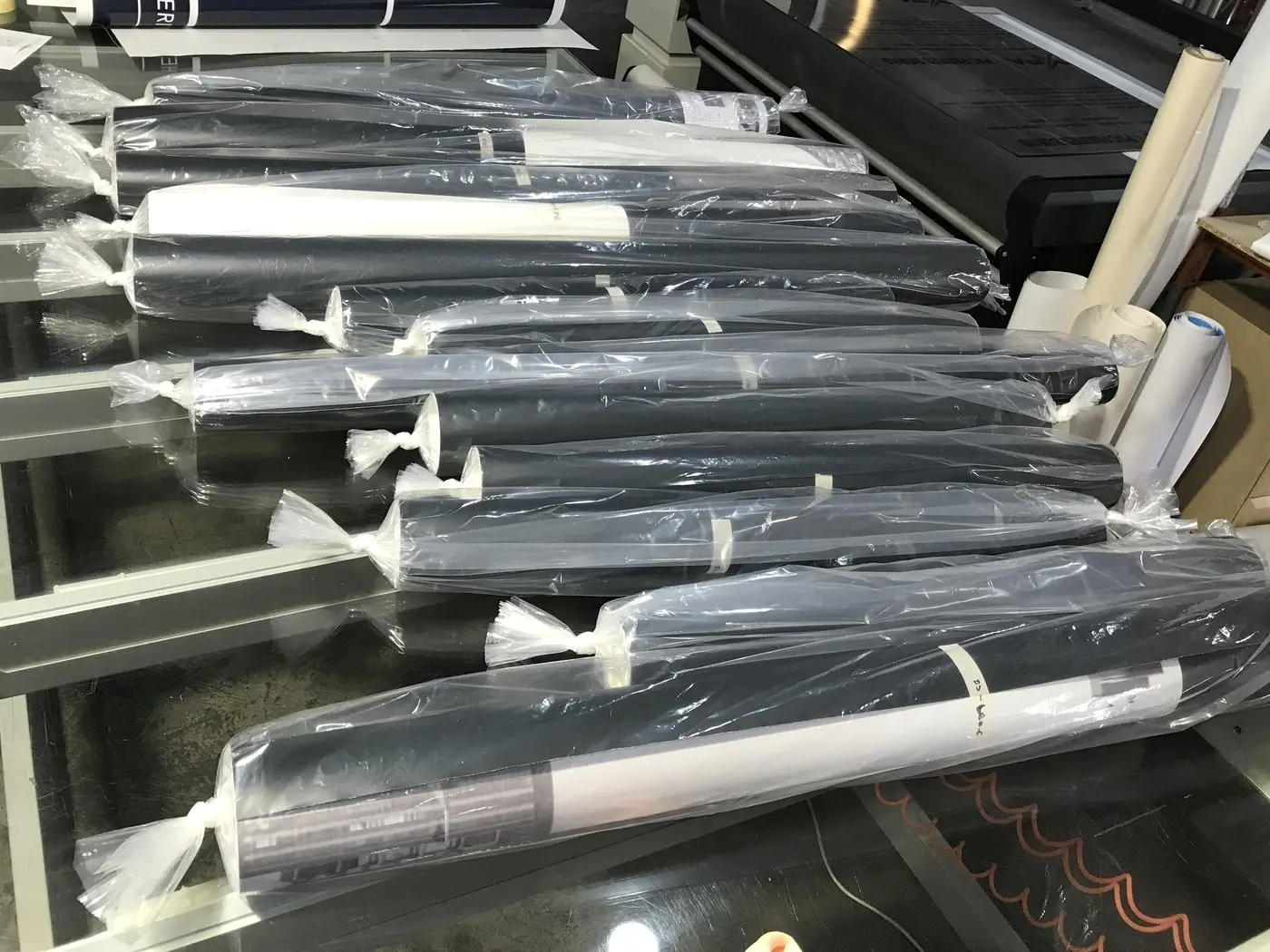 Nine rolls of printed black hoarding graphics wrapped in plastic on the workshop bench beside the laminator