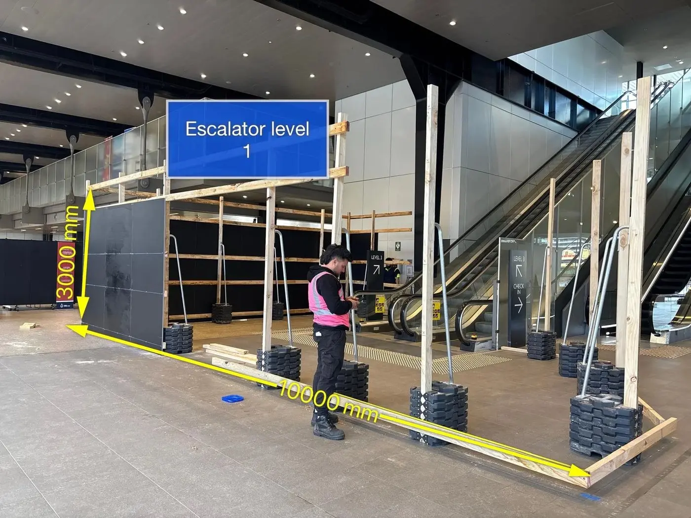 Installer measuring a hoarding frame beside an escalator with 3000mm and 10000mm dimension overlays marked on the photograph
