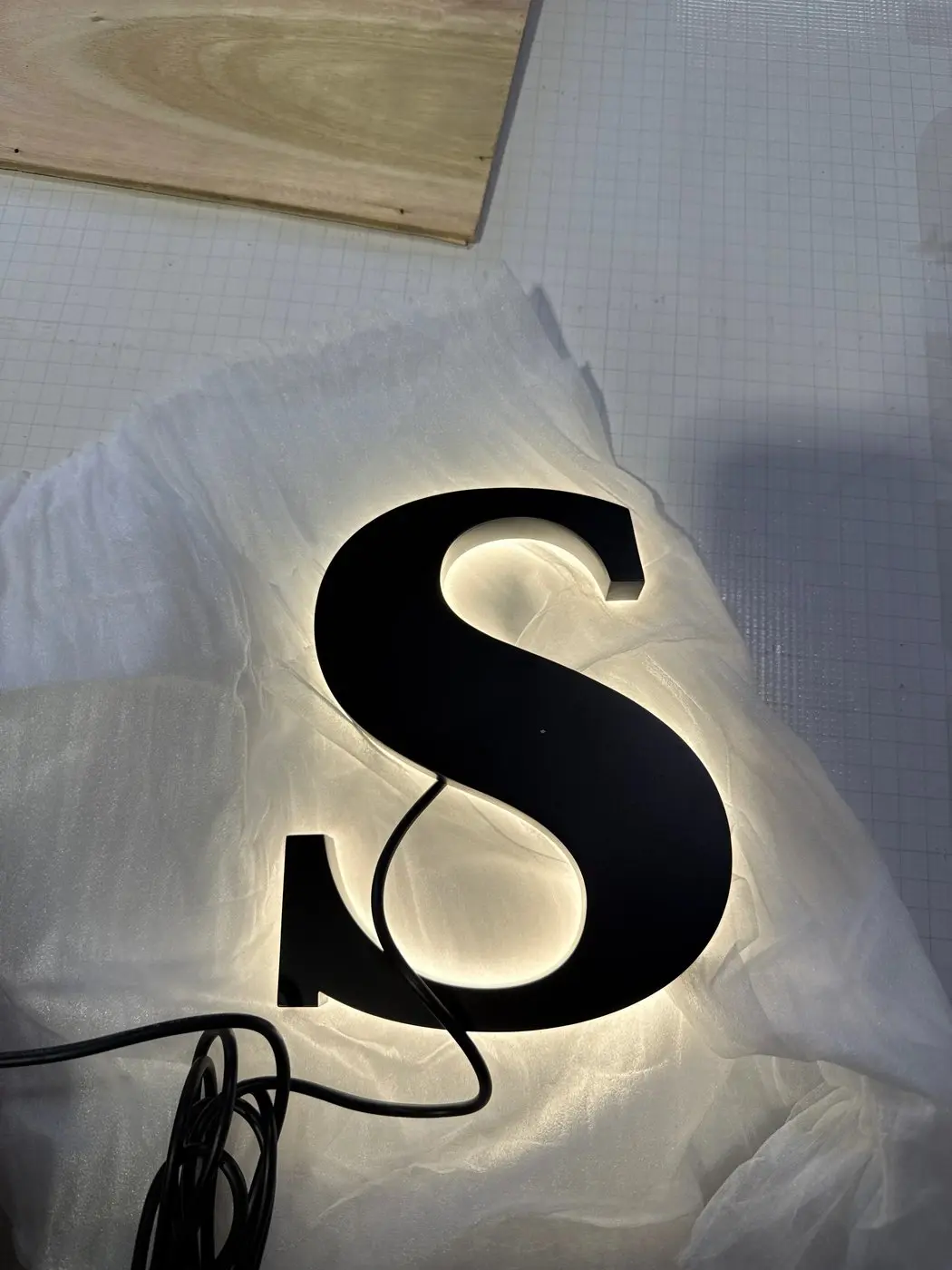 Single matt-black letter S powered up during quality checks with its halo glow visible against the packing sheet