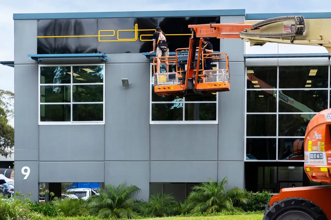 Installers aligning the final section of black ACM beside the yellow dimensional EDA logo
