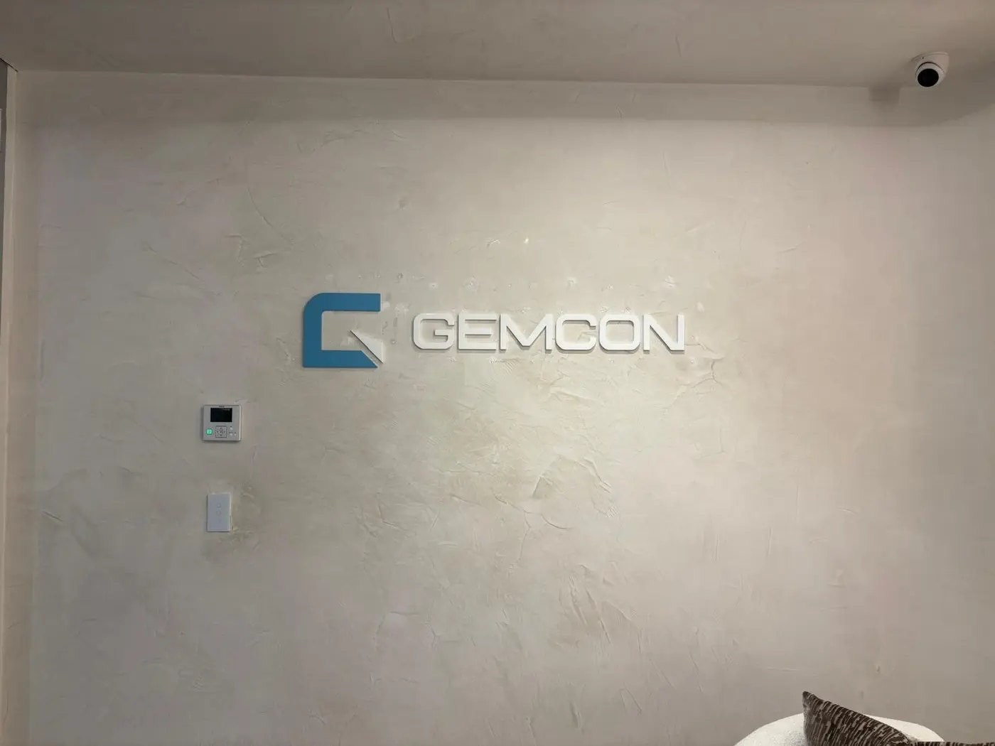 White dimensional GEMCON lettering with a painted blue logo icon mounted on a textured plaster reception wall