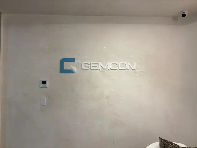 White dimensional GEMCON lettering with a painted blue logo icon mounted on a textured plaster reception wall