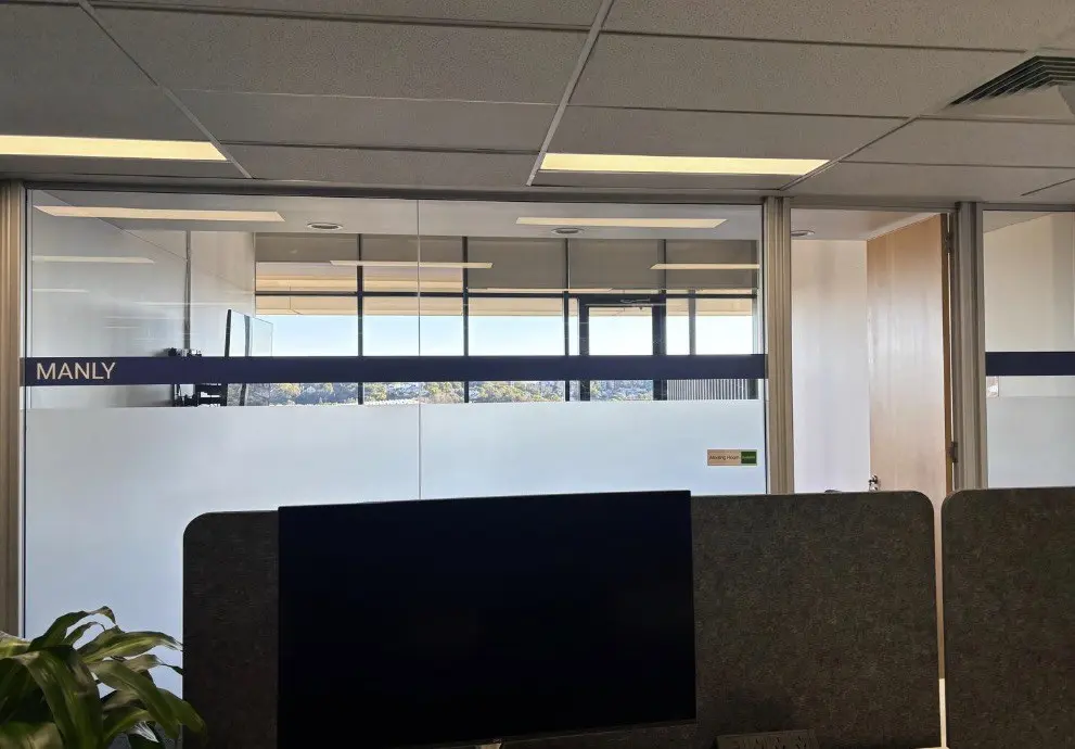 Meeting room named Manly applied as cut vinyl lettering to the dark band of a glazed office partition with harbour light behind
