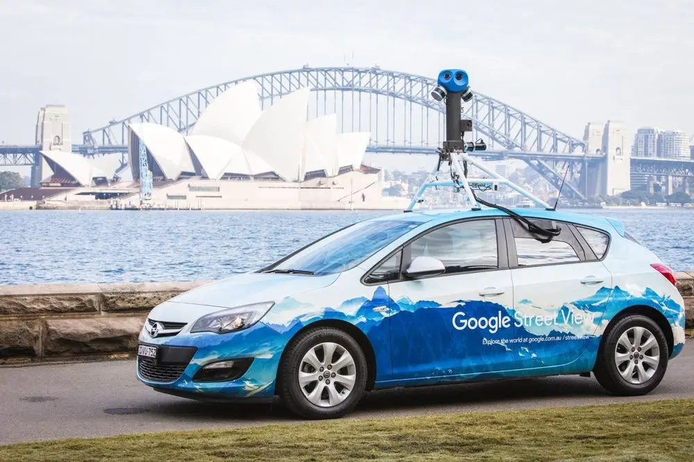 Google Street View car in its printed blue livery with roof camera rig, photographed in front of the Sydney Opera House and Harbour Bridge