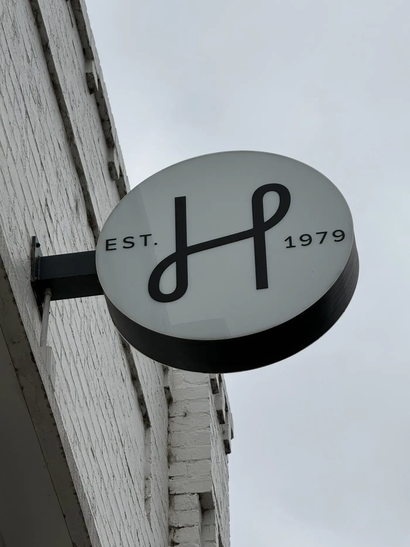 Round double-sided LED light box blade sign carrying the HALI H monogram and EST 1979, mounted on a painted brick wall against the sky