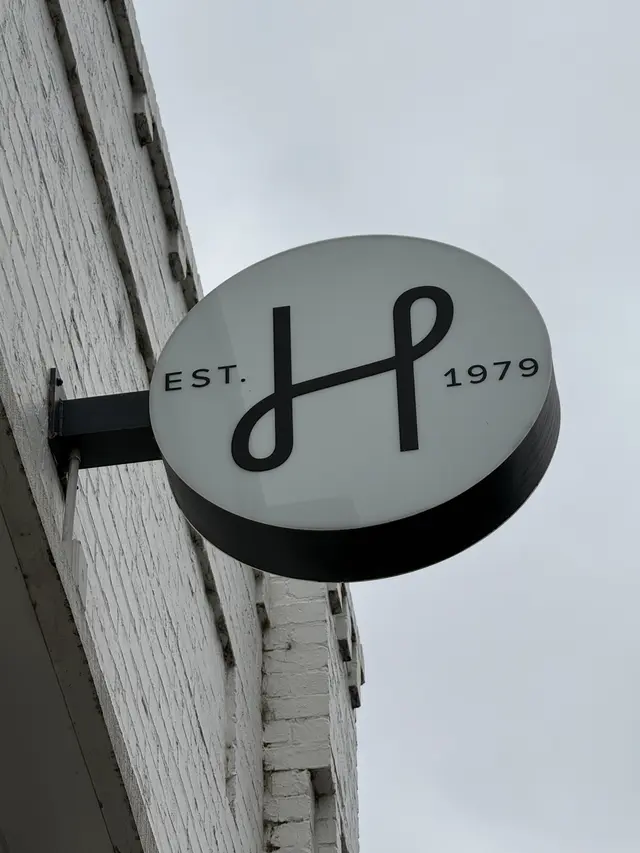 Round double-sided LED light box blade sign carrying the HALI H monogram and EST 1979, mounted on a painted brick wall against the sky