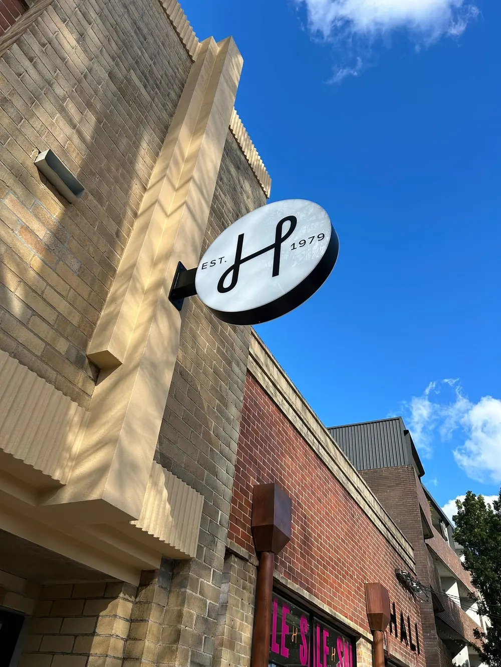 Round double-sided LED light box with the HALI H monogram projecting from a sandstone corner pier against a blue sky