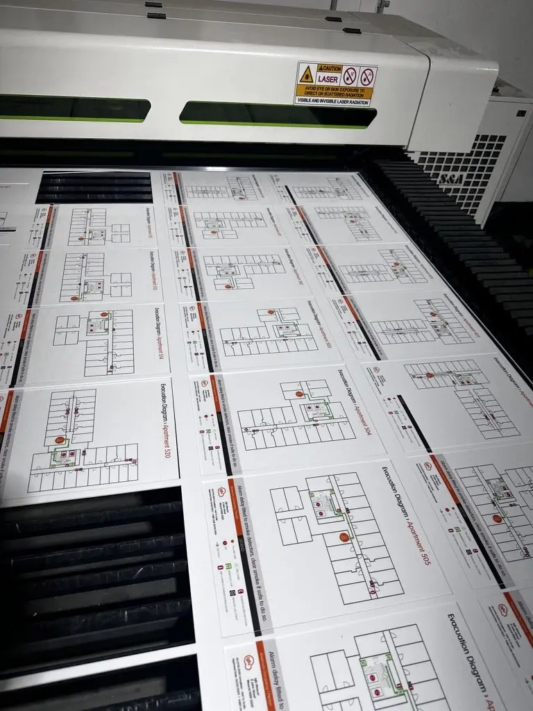 Printed apartment-specific evacuation diagram panels nested on the laser cutting bed, each labelled with a different apartment number