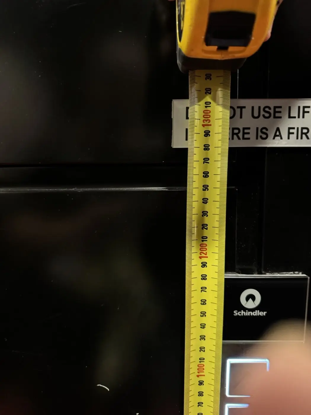Tape measure held against a lift sign to check its measured mounting height beside the lift controls