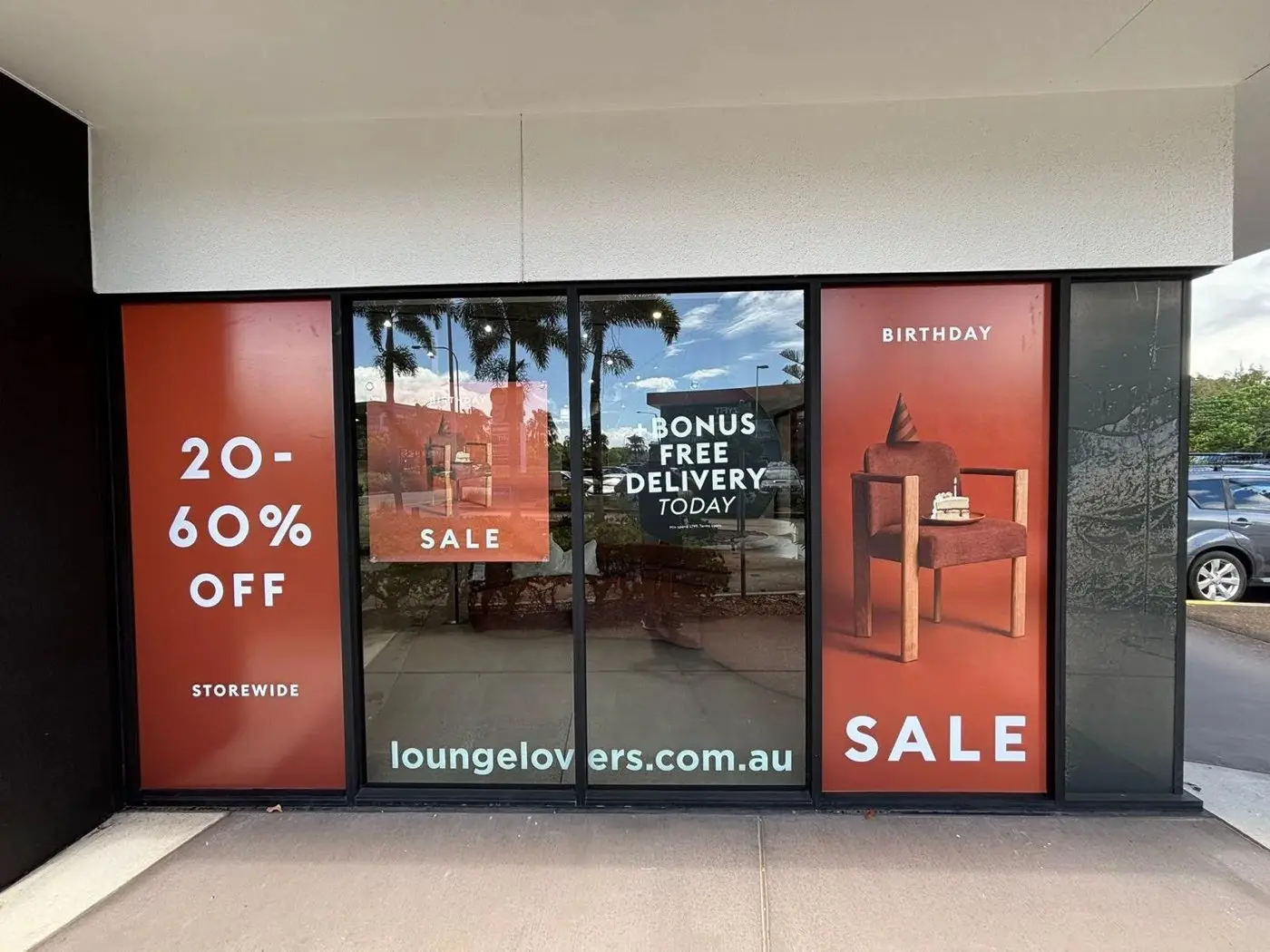 Showroom windows with Birthday Sale and free delivery panels, palm trees reflected in the glass