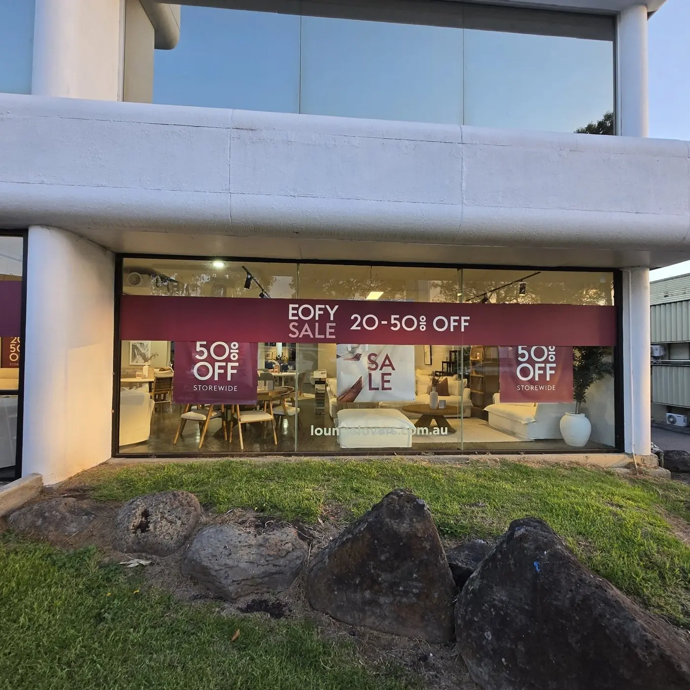 A second Lounge Lovers showroom at dusk with EOFY sale and 50 percent off window graphics lit from inside