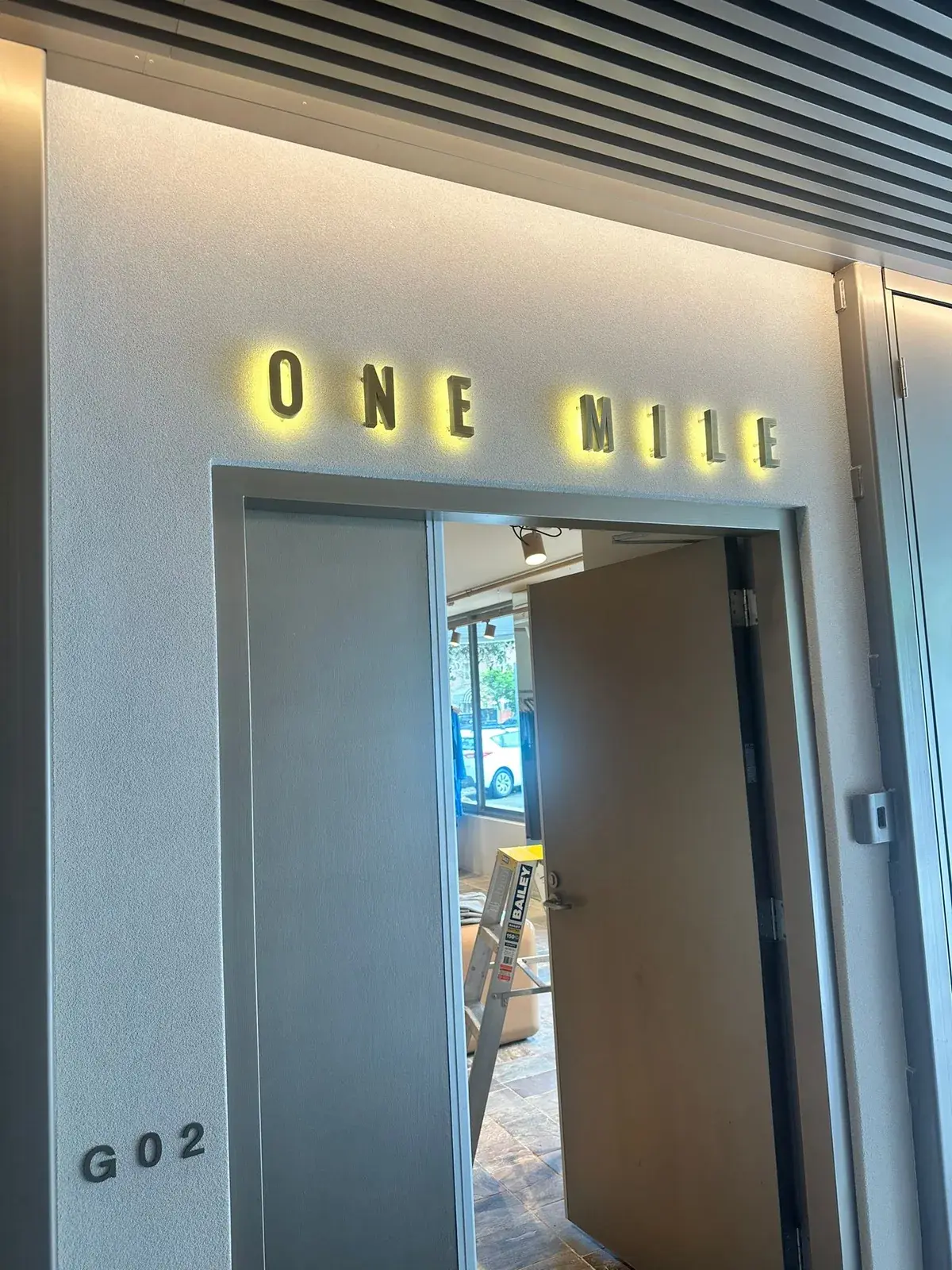 Angled view of the illuminated ONE MILE entry lettering and warm halo against the textured boutique wall
