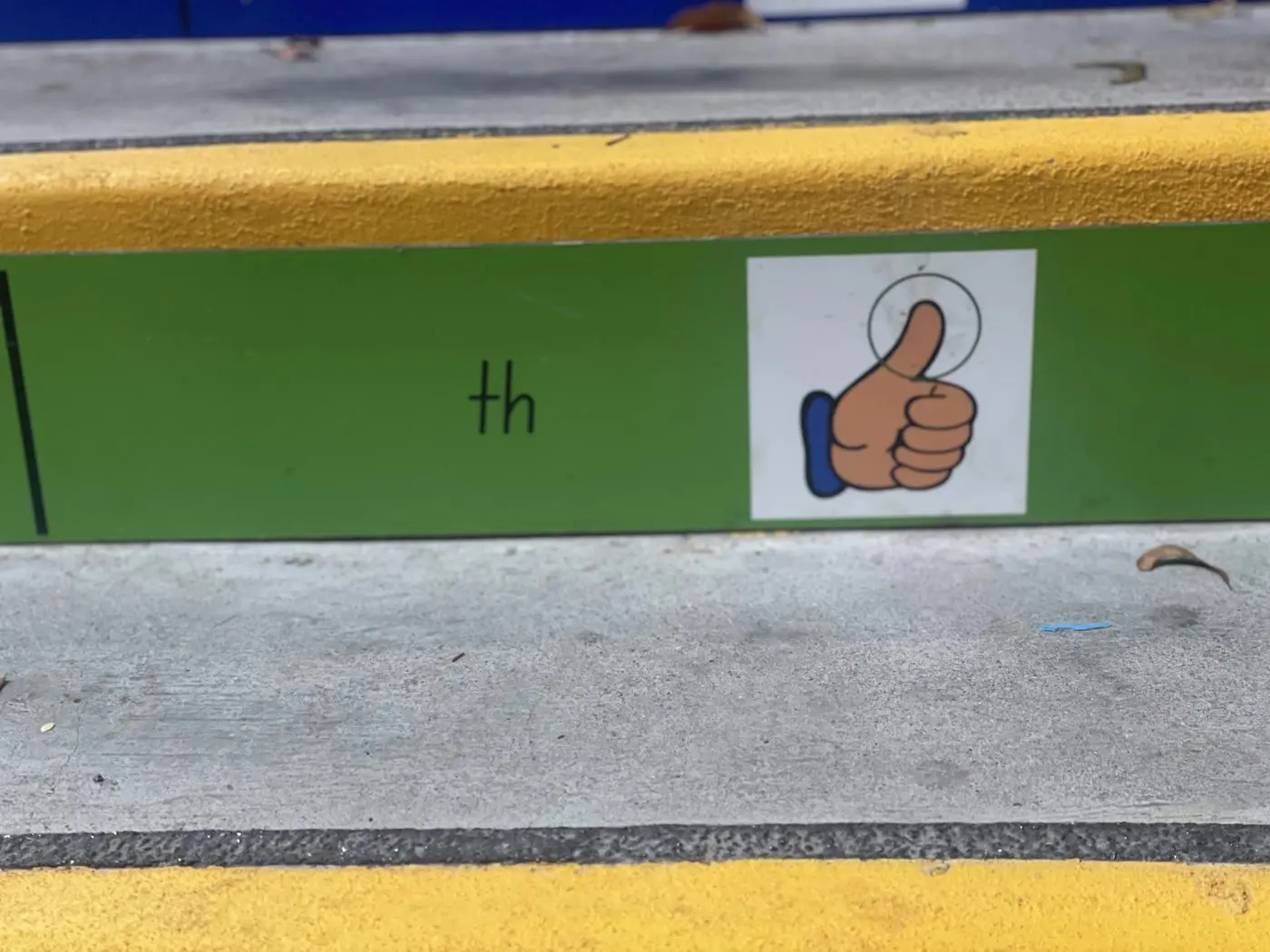 Close view of a green playground wall band pairing the phonics sound th with a thumbs up picture