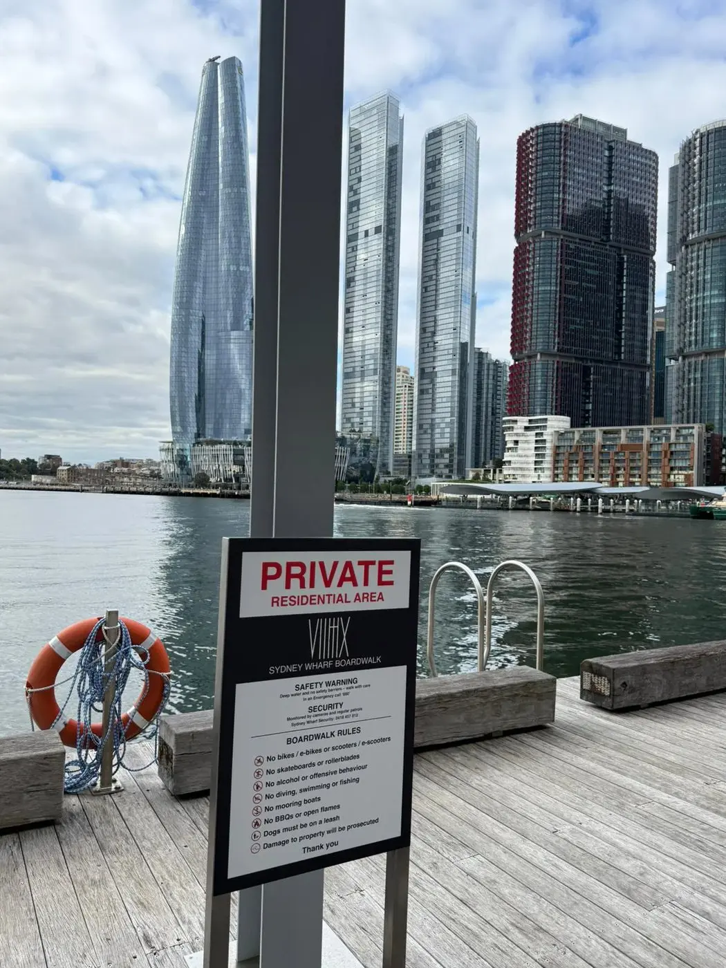 Main regulatory sign mounted on a pole at the Sydney Wharf Apartments boardwalk with the harbour and Barangaroo towers behind