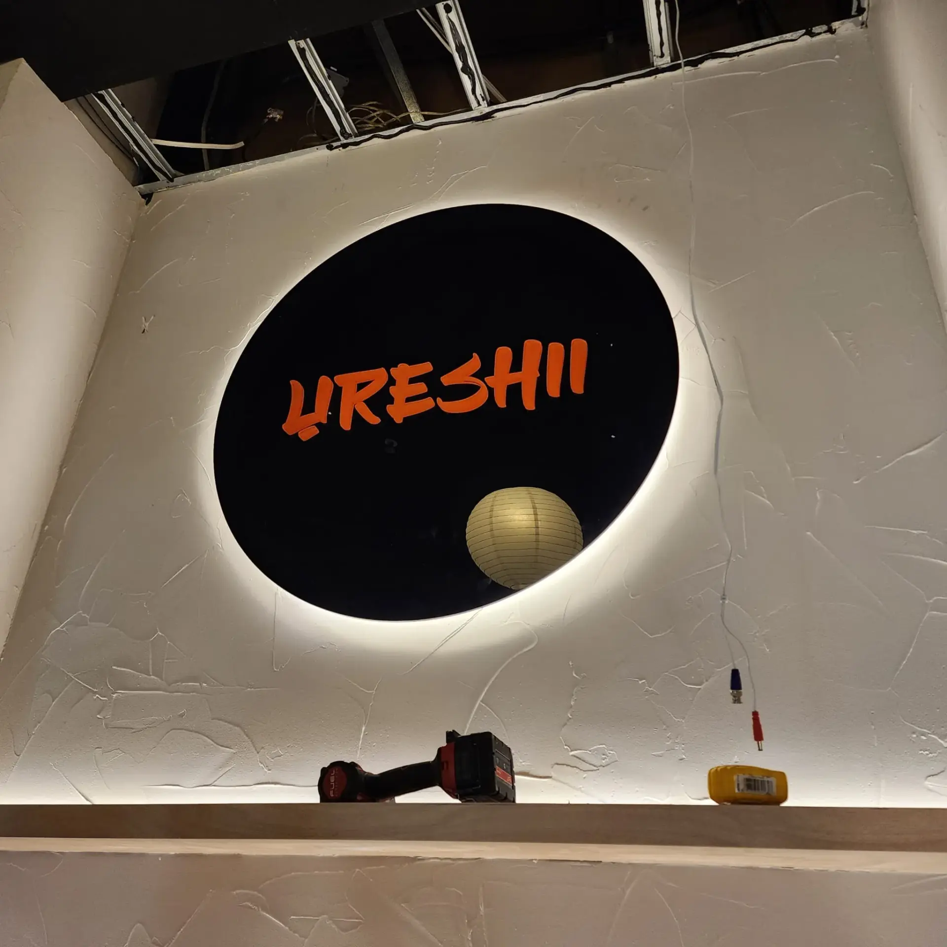 Round black Ureshii interior feature sign with orange lettering and a white halo glow