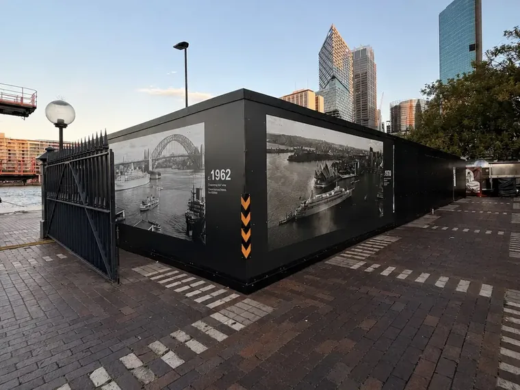 Site compound hoarding on the Circular Quay forecourt wrapped around its corner with c.1962 and 1970 archival harbour photographs, heritage gates alongside and the city skyline behind