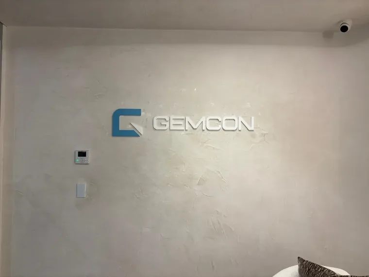 White dimensional GEMCON lettering with a painted blue logo icon mounted on a textured plaster reception wall
